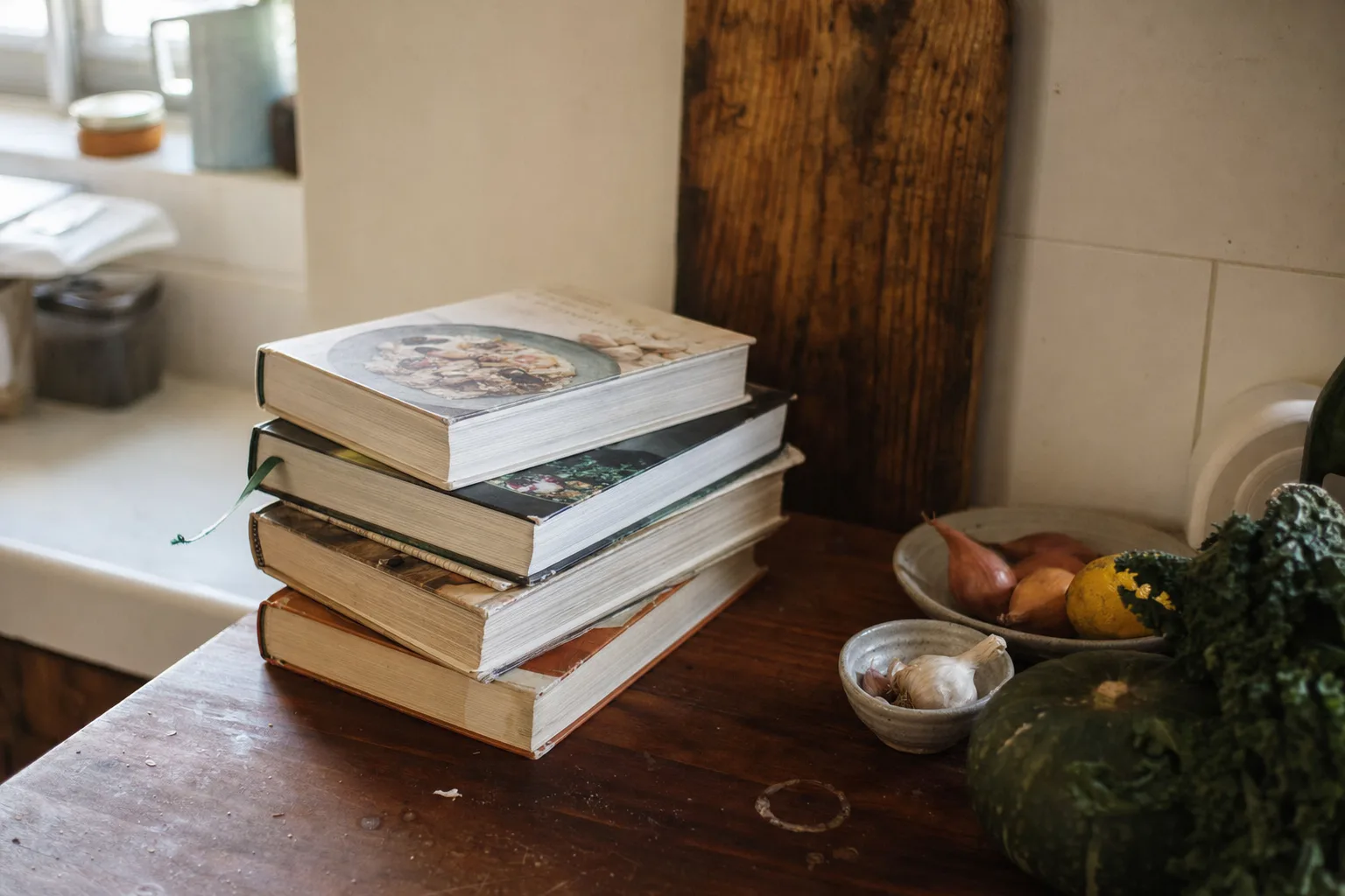 A stack of worn cookbooks on a kitchen counter with a bookmark sticking out and ingredients pushed aside