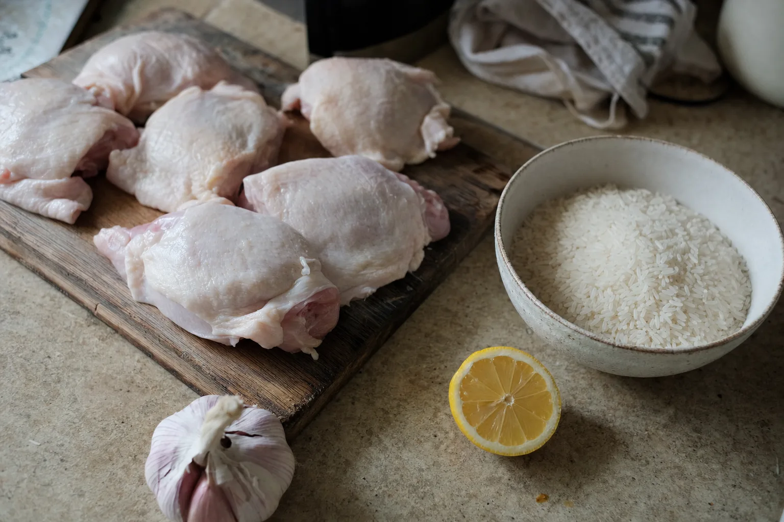 Raw chicken thighs on a wooden board next to a bowl of uncooked white rice, garlic and half a lemon