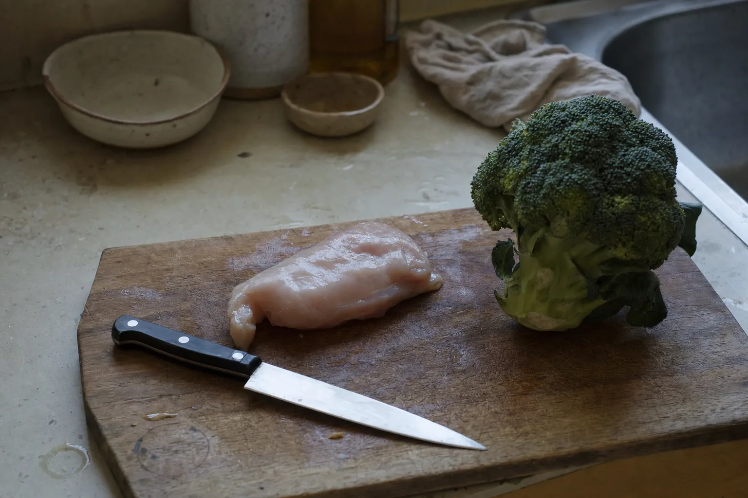 A raw chicken breast and a head of broccoli on a scarred cutting board with a chef knife