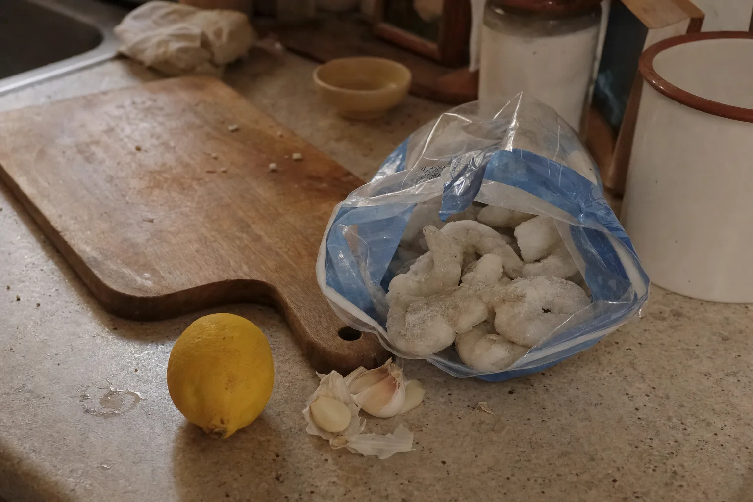 An opened bag of frozen shrimp on a counter with ice crystals still on them, lemon and garlic nearby