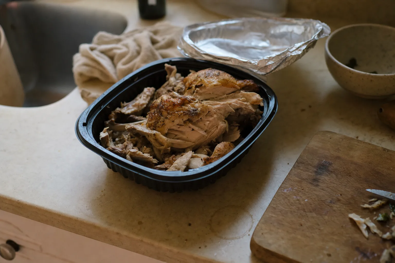 A picked-over rotisserie chicken in its supermarket container with the foil lid peeled back