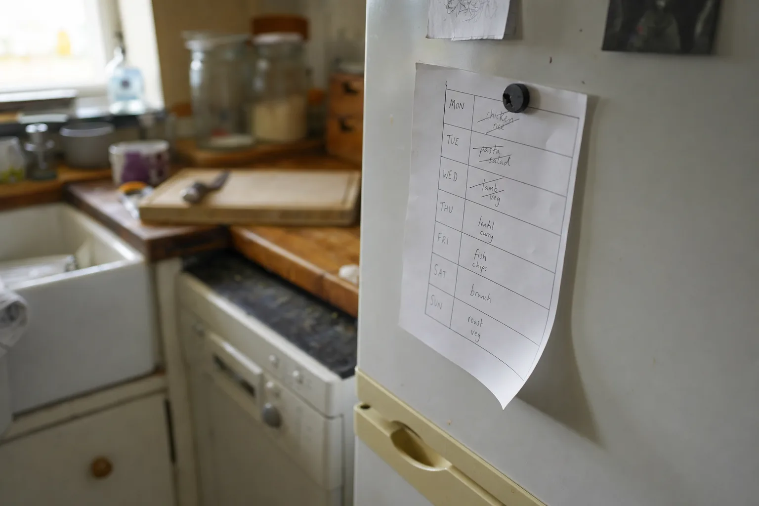 A paper weekly meal plan held to a fridge door by a magnet, curling at one corner with days crossed out