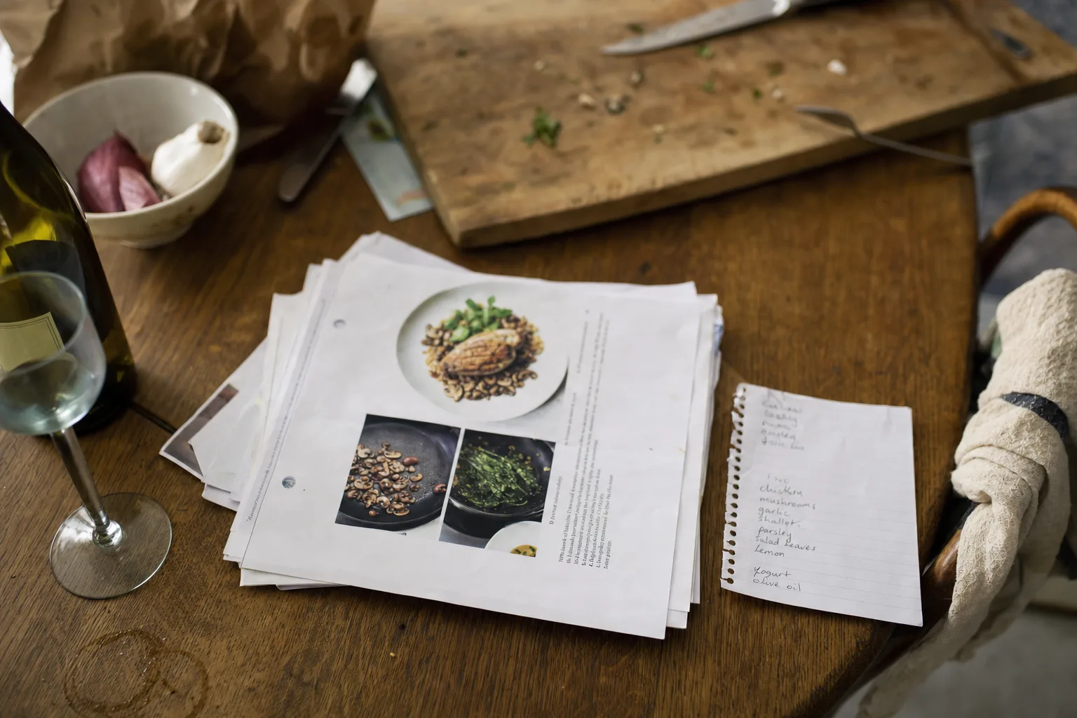 A stack of printed recipe pages and a torn-off handwritten shopping list on a kitchen table