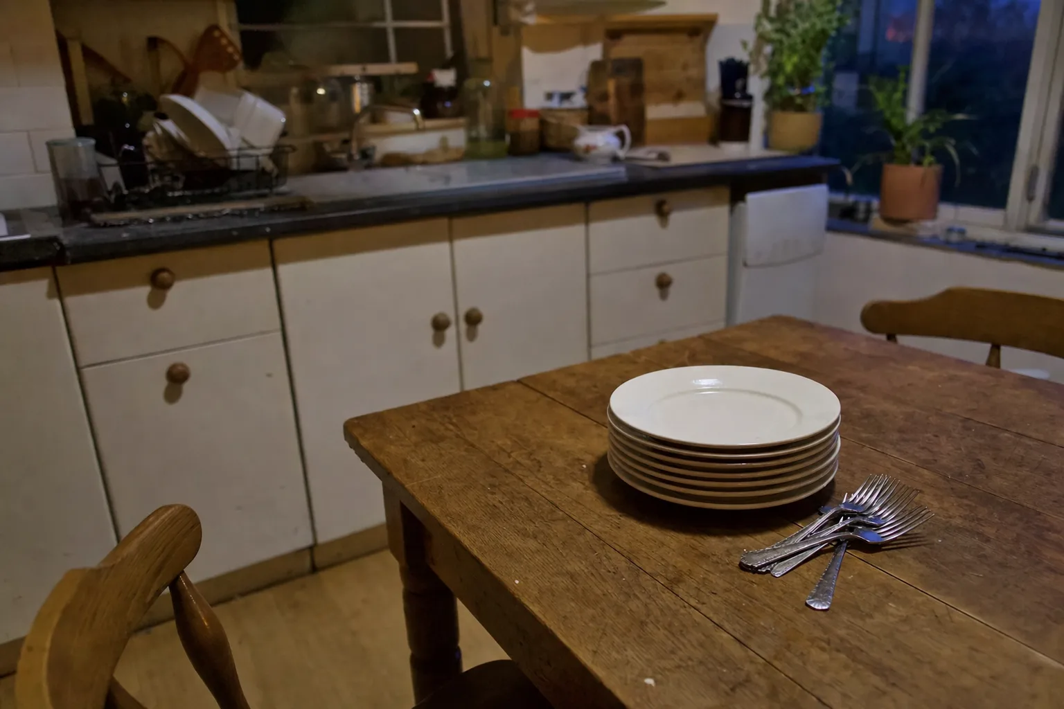 A stack of clean dinner plates and forks at the end of an empty kitchen table at dusk
